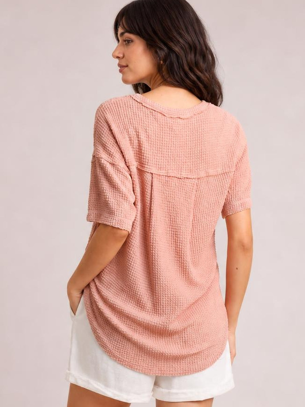 A model turns to show the relaxed silhouette of a pink waffle-knit half-sleeve T-shirt with a rounded hem and textured back construction.