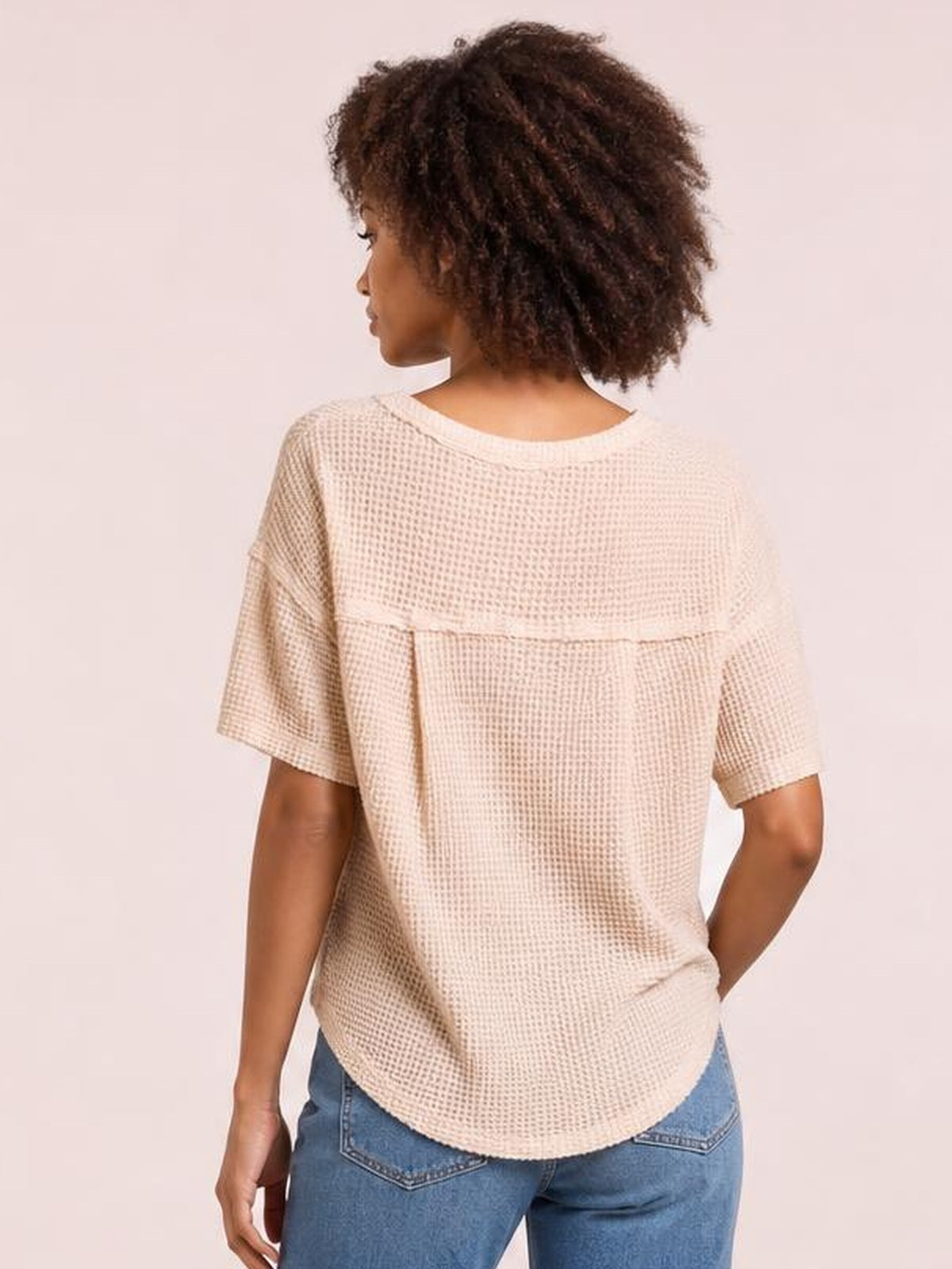 Model shown from the back wearing a cream waffle-knit half-sleeve T-shirt with a notched neckline, yoke seam, relaxed silhouette, and curved hem.