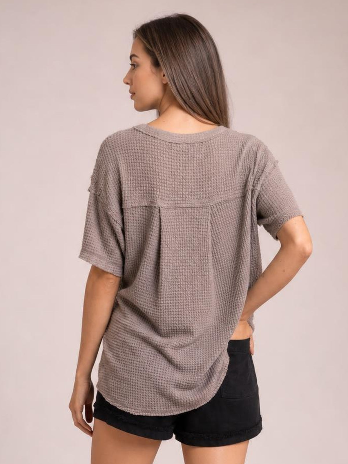 Model shown from behind wearing a taupe waffle-knit T-shirt with relaxed silhouette, loose half sleeves, curved hem, and a large back pocket.