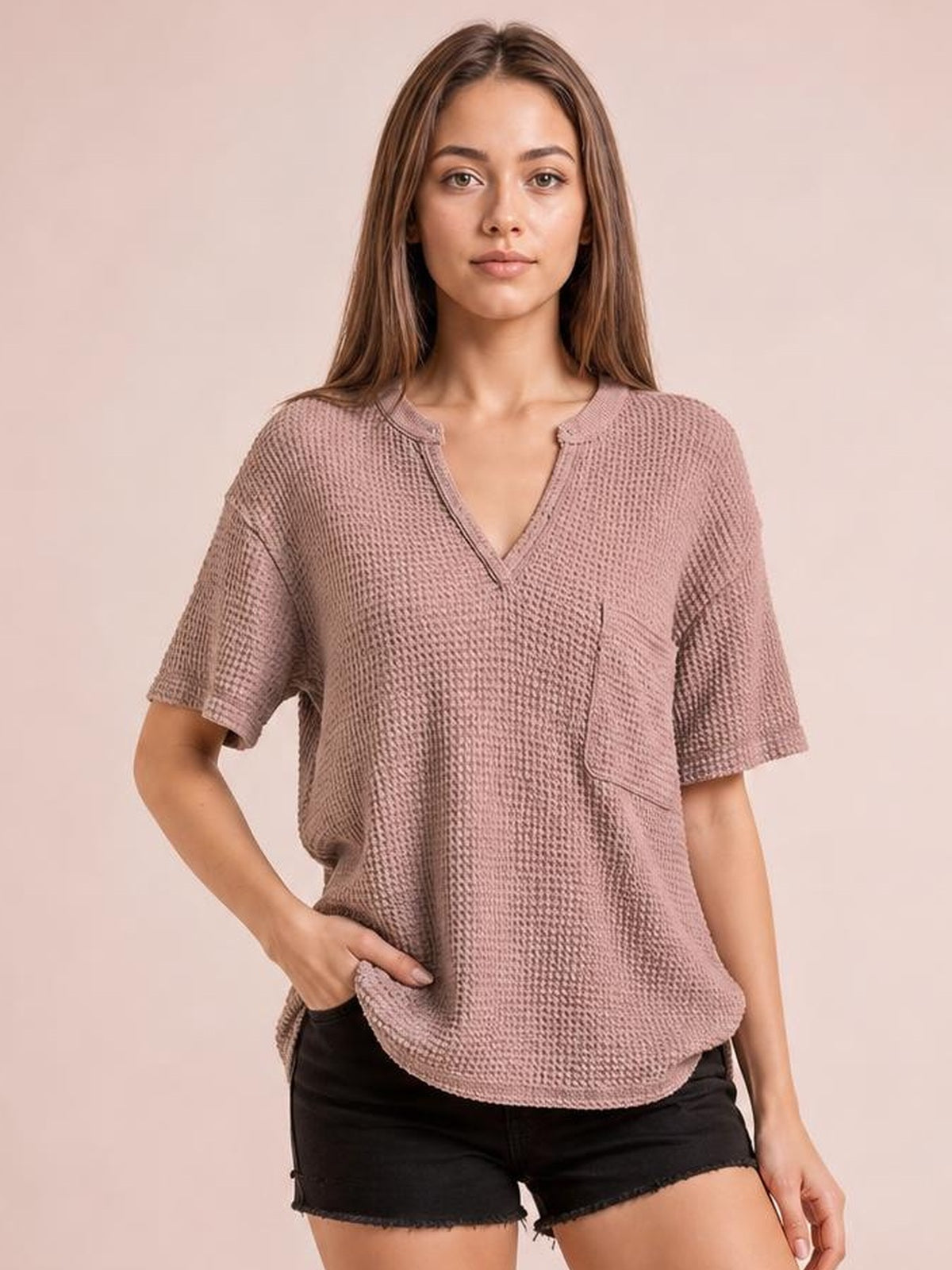 Model wears a mauve waffle-knit T-shirt with a notched neckline, half sleeves, relaxed silhouette, curved hem, and front patch pocket, posing with one hand in it.