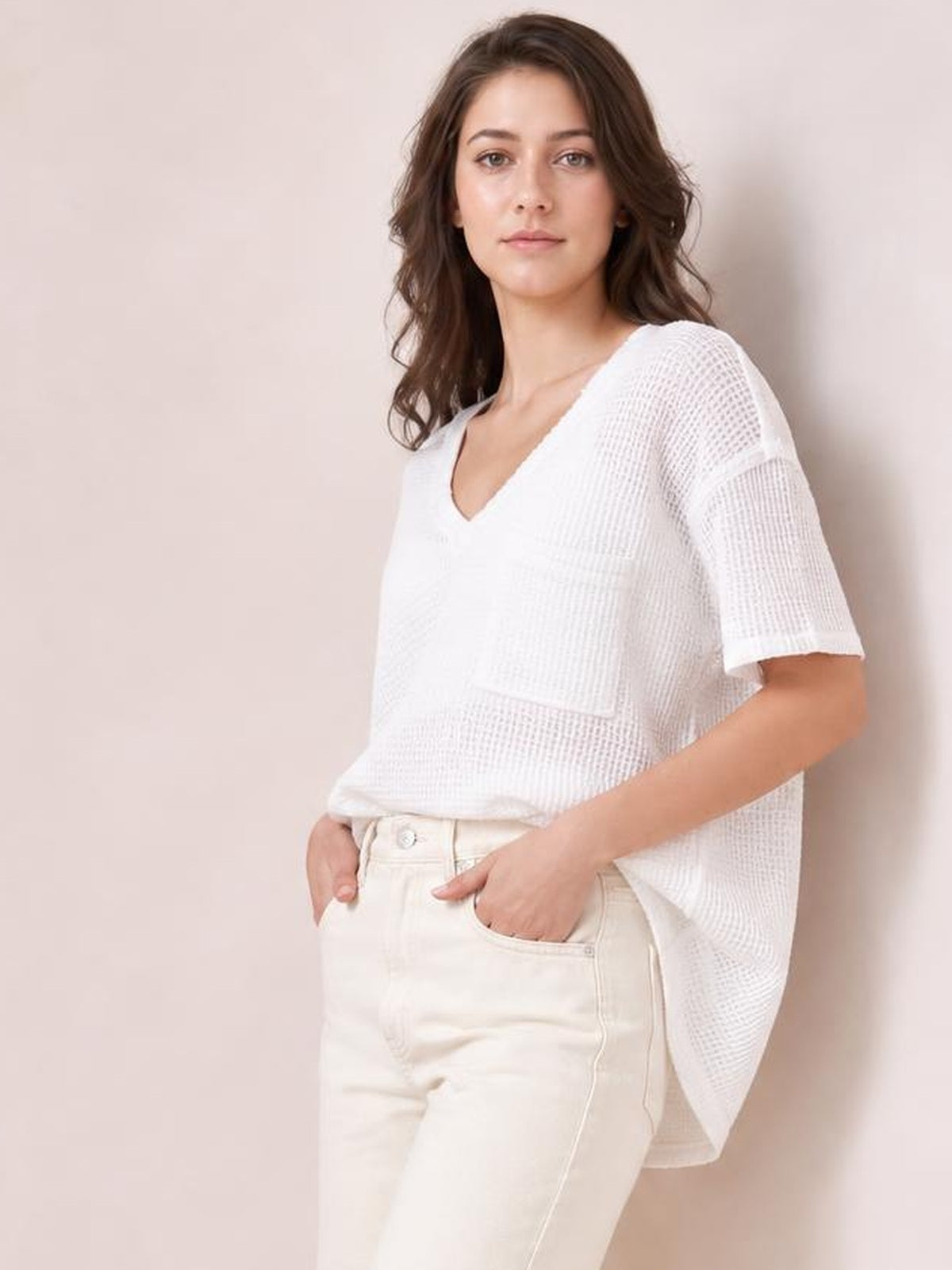A model poses in a white waffle-knit T-shirt with a notched V-neck, half sleeves, and a relaxed high-low silhouette, hands in her jeans pockets.