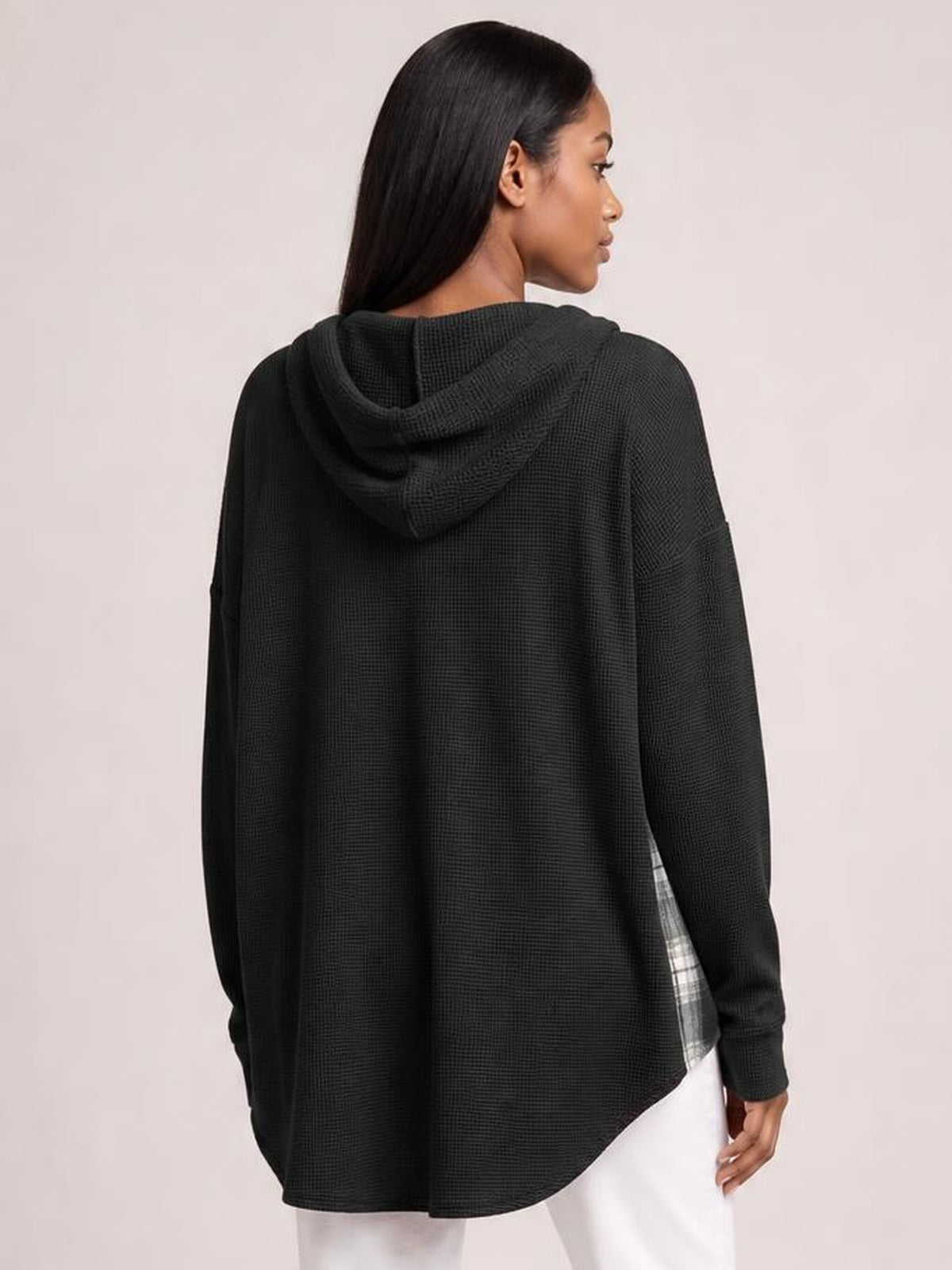 A model turns to show a black waffle knit hooded shacket with a relaxed oversized silhouette, long sleeves, curved hem, and subtle plaid patchwork at the side.