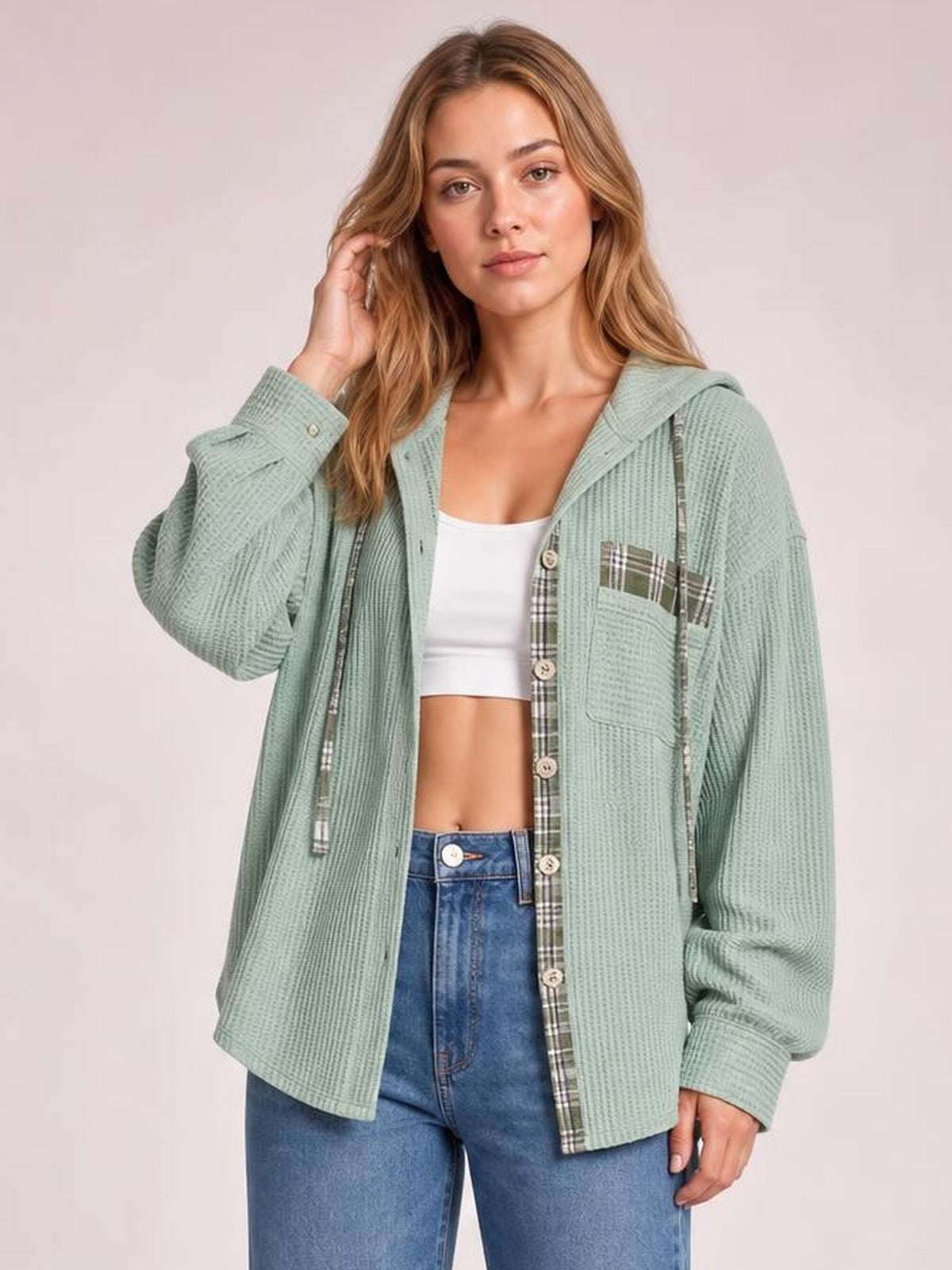 A woman poses in a sage green waffle knit hooded shacket with plaid patchwork trim, drawstring hood, button front, pockets, and a relaxed silhouette.