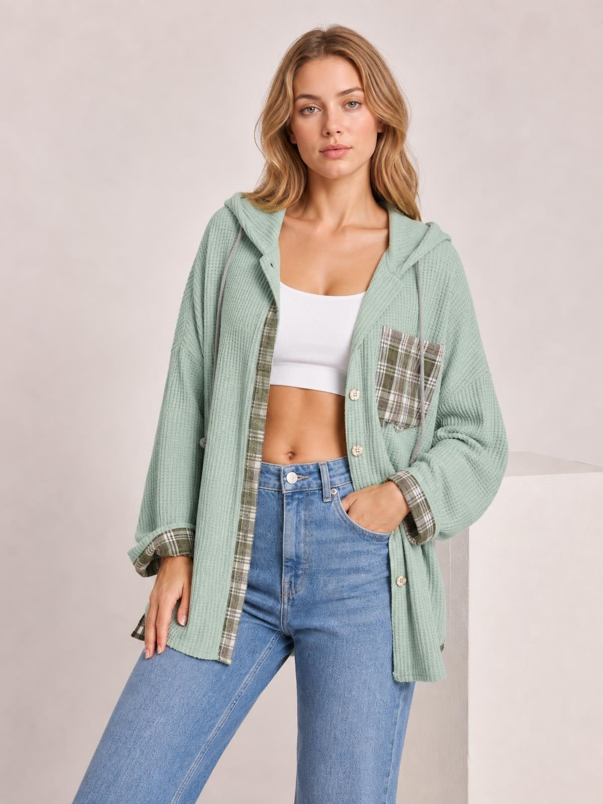 A model stands in a sage green waffle-knit hooded shacket with plaid patchwork trim, button front, chest pocket, and a relaxed long silhouette.