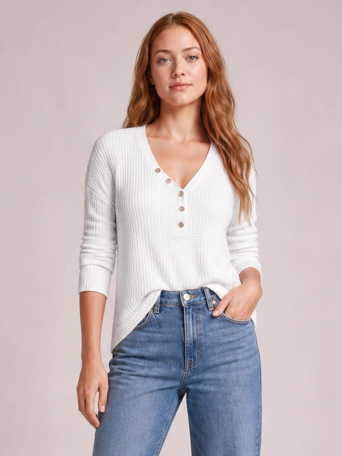 Model wears an opaque white waffle-knit V-neck long-sleeve top with a relaxed silhouette and button details, posing with one hand in her jeans pocket.