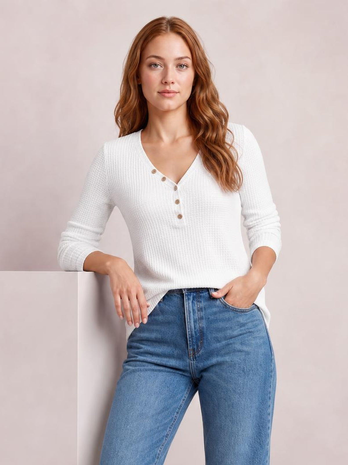 A woman wears a white waffle-knit V-neck long sleeve top with button details and a relaxed straight silhouette, posing with one hand in her jeans pocket.