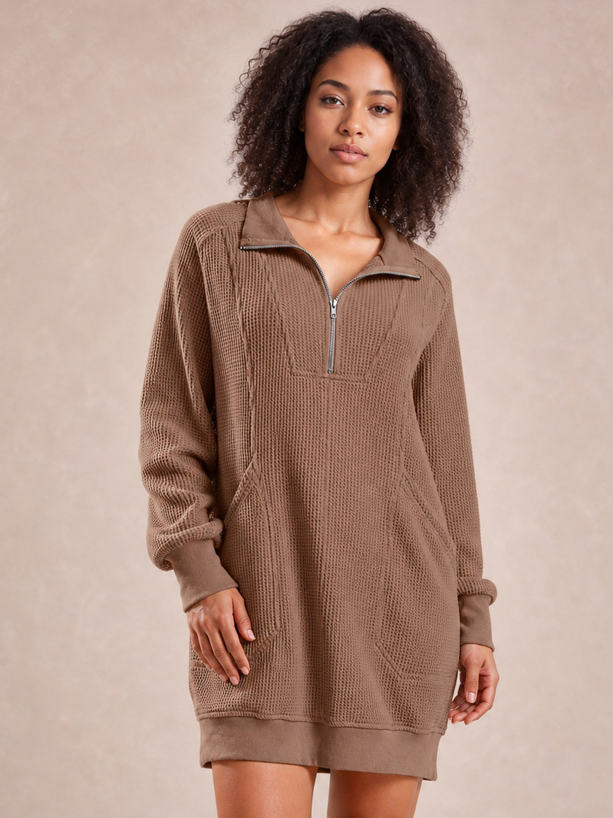 Model wears a taupe waffle-knit mini dress with long sleeves, a relaxed silhouette, half-zip collar, ribbed cuffs and hem, and side pockets.