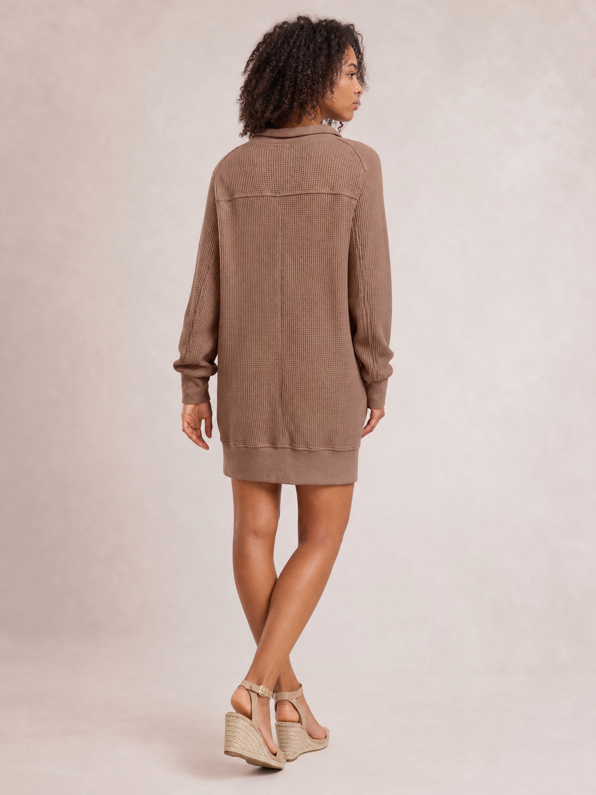 Model looks over her shoulder in a taupe waffle-knit long-sleeve mini dress with a relaxed straight silhouette, textured panels, and ribbed cuffs and hem.