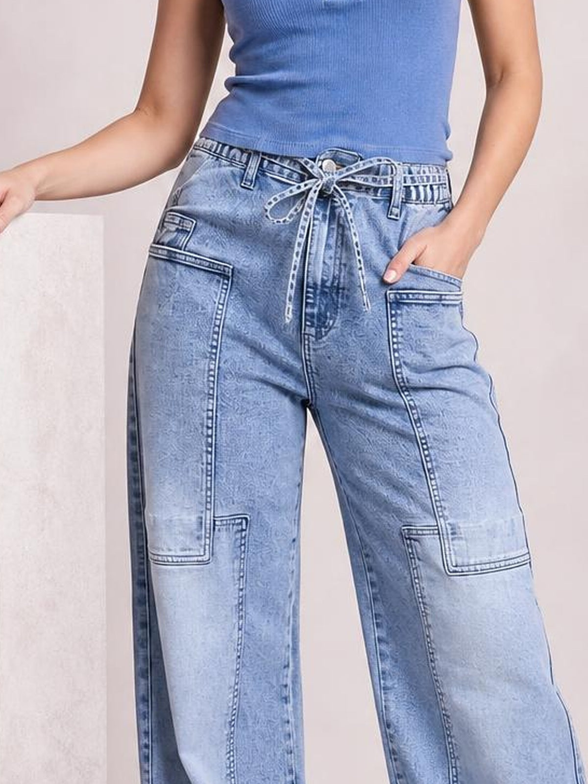 A model wears light-wash wide-leg cargo jeans with a tied waist, multiple patch pockets, and prominent contrast stitching, posing with one hand in a pocket.