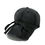Washed dark denim baseball cap with a curved brim, seamed crown, top button, ventilation eyelets, and an oversized front bow with trailing ends.