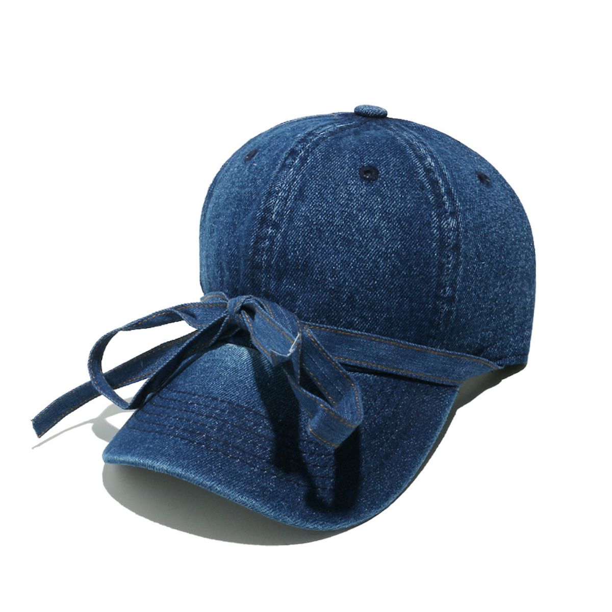 Washed blue denim baseball cap with a seamed crown, curved brim, top button, ventilation eyelets, and an oversized front bow with trailing ends.