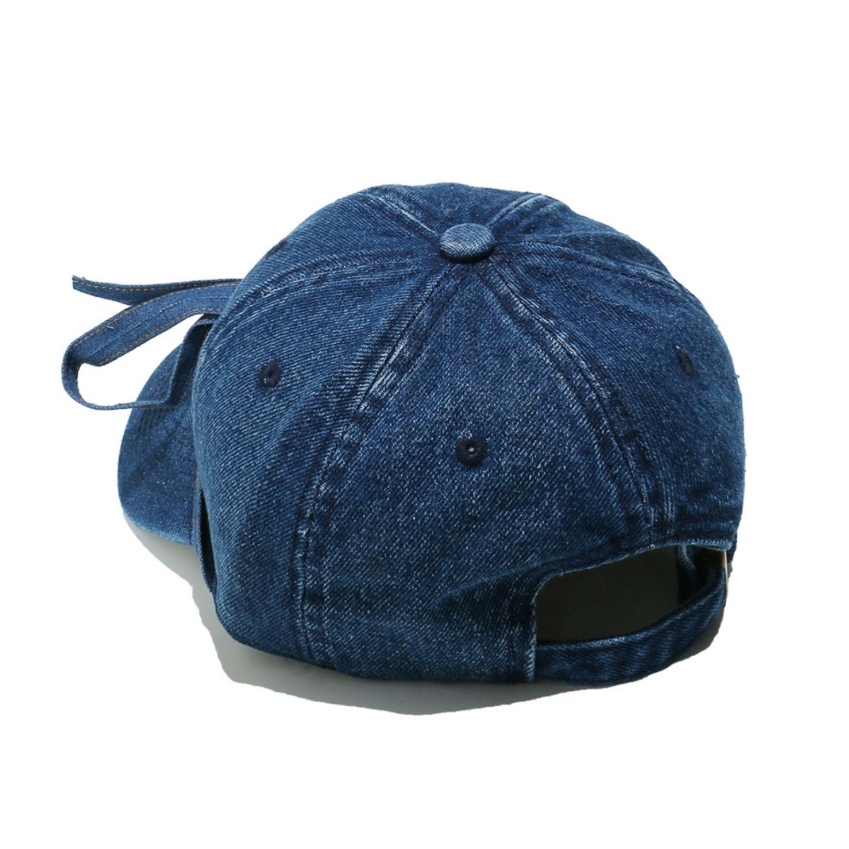A washed blue denim baseball cap features a seamed crown, top button, ventilation eyelets, curved brim, adjustable back opening, and trailing bow detail.