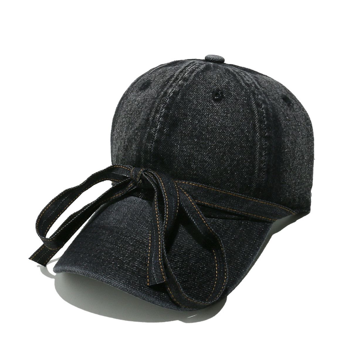 Washed dark denim baseball cap with a curved brim, seamed crown, top button, ventilation eyelets, and an oversized front bow with trailing ends.