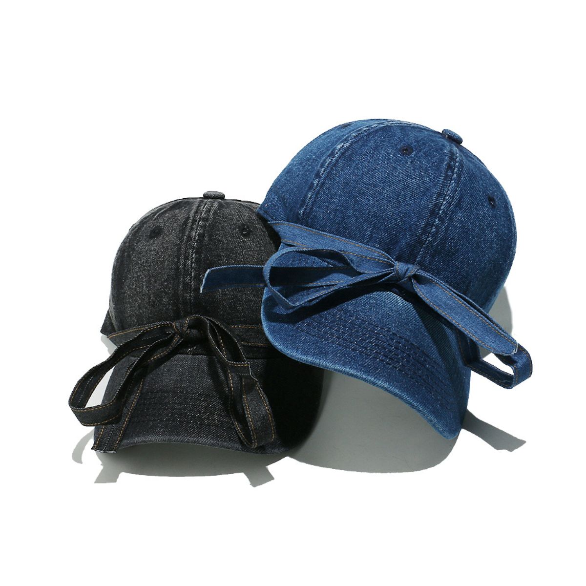 Washed denim baseball caps in blue and black feature seamed crowns, curved brims, top buttons, ventilation eyelets, and oversized front bows with trailing ends.