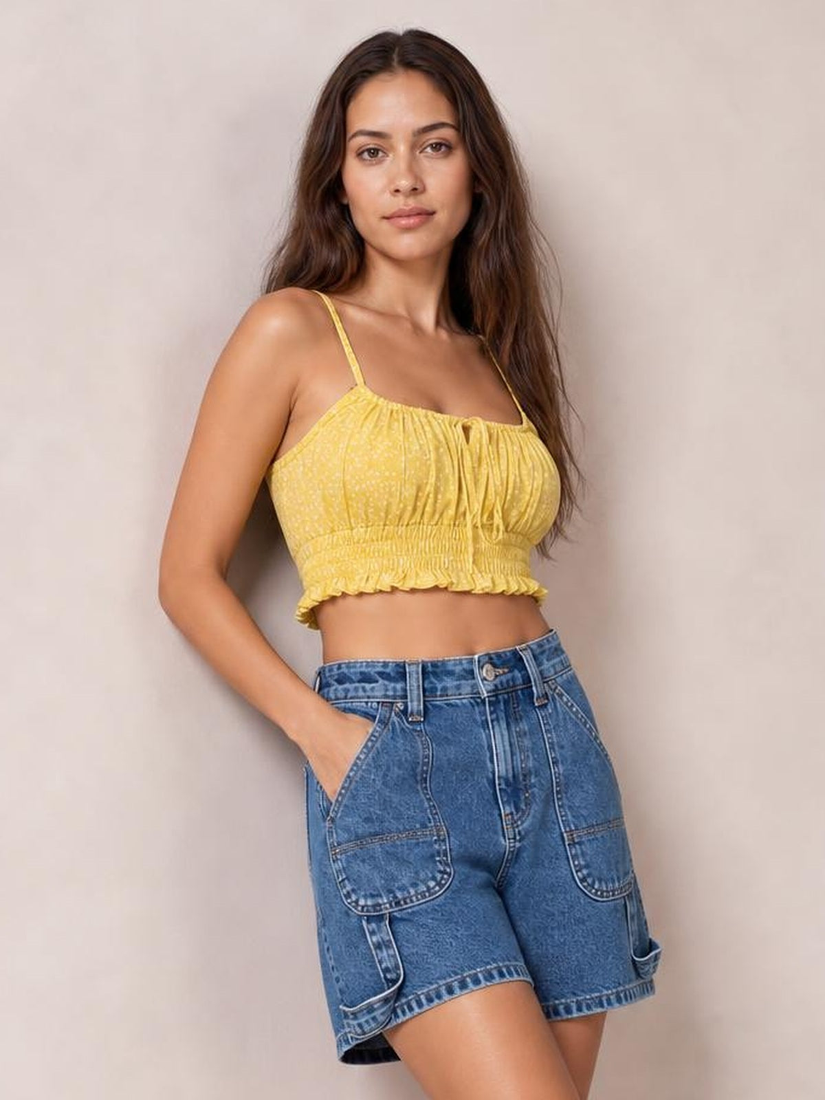 A woman modeling a yellow textured smocked crop top with thin spaghetti straps and a ruffled hem, paired with blue denim shorts, against a neutral beige background.