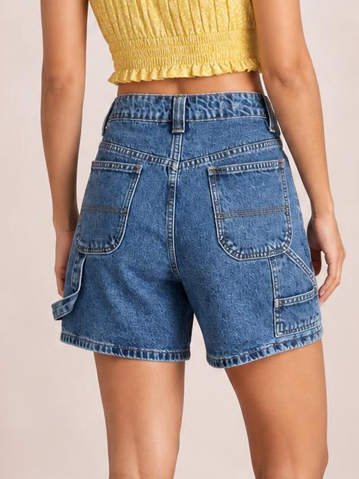 A model stands with her back to the camera wearing washed blue high-rise denim shorts with belt loops, contrast stitching, large back pockets, and side cargo pockets.