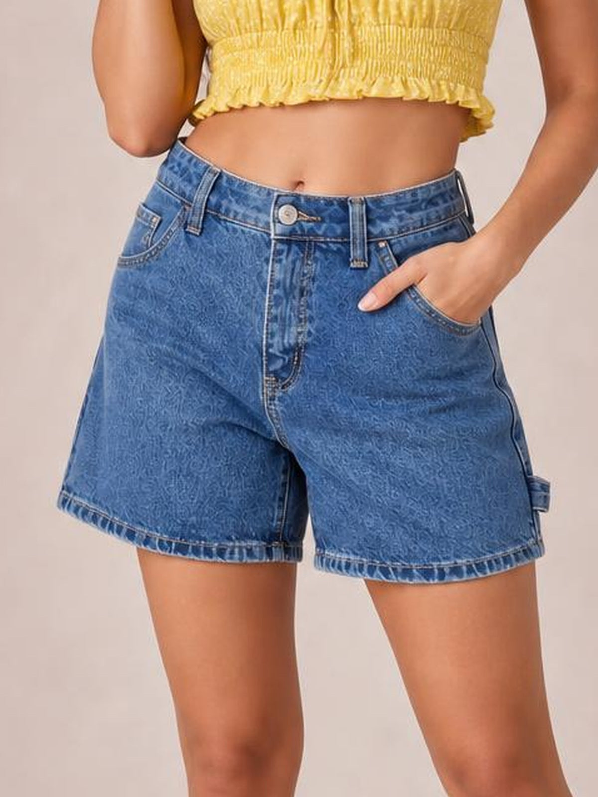 A model wears washed high-rise denim shorts in blue with a relaxed silhouette, belt loops, button closure, front pockets, and stitched hems, one hand tucked in a pocket.