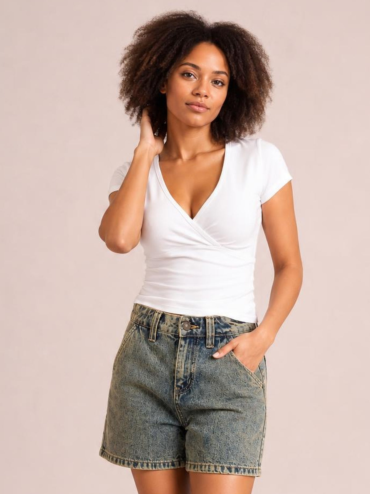 A woman modeling a white wrap-style short-sleeve V-neck top and high-waisted blue denim shorts against a solid light beige studio background.