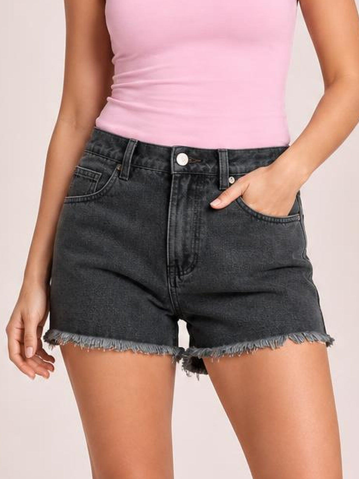A model wears washed black denim shorts with a high-rise silhouette, belt loops, front pockets, button closure, and frayed raw hems, posing with one hand in a pocket.