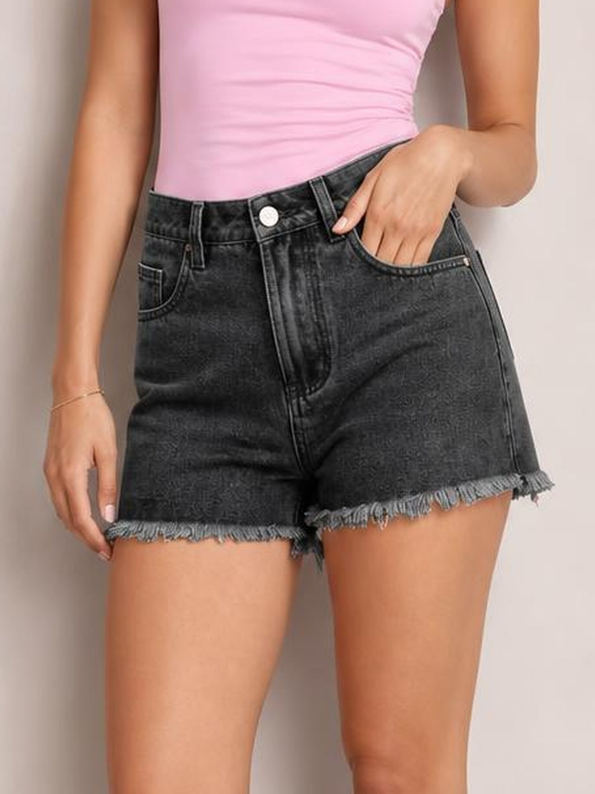 Model wearing washed black denim shorts with a high-rise button fly, belt loops, front pockets, relaxed silhouette, and frayed raw hems, one hand in a pocket.
