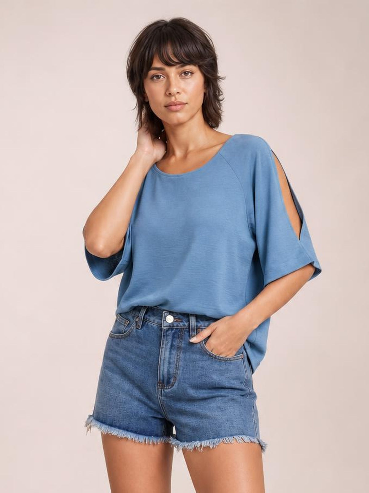 Model wearing blue washed denim shorts with a high-rise silhouette, front pockets, button closure, belt loops, and frayed raw hems, with one hand in a pocket.
