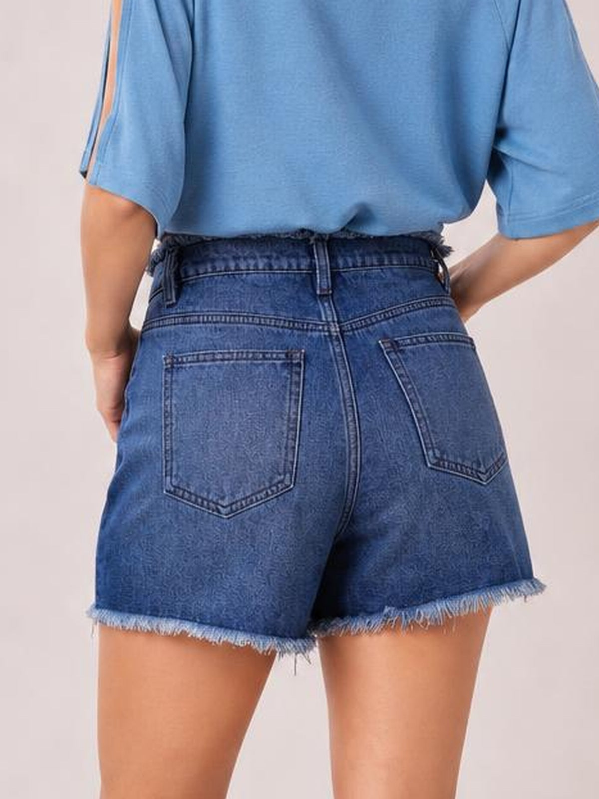 Model shown from the back stands with hands at her hips in washed blue denim shorts featuring a high waistband, back pockets, and frayed raw hems.