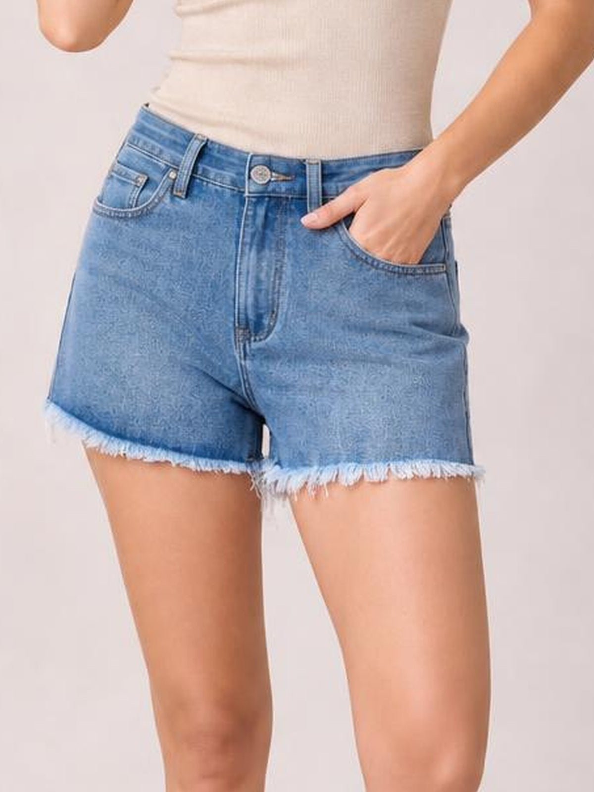 Model wearing light blue washed denim shorts with a relaxed silhouette, raw frayed hems, belt loops, button fly, and front pockets, with one hand tucked inside.