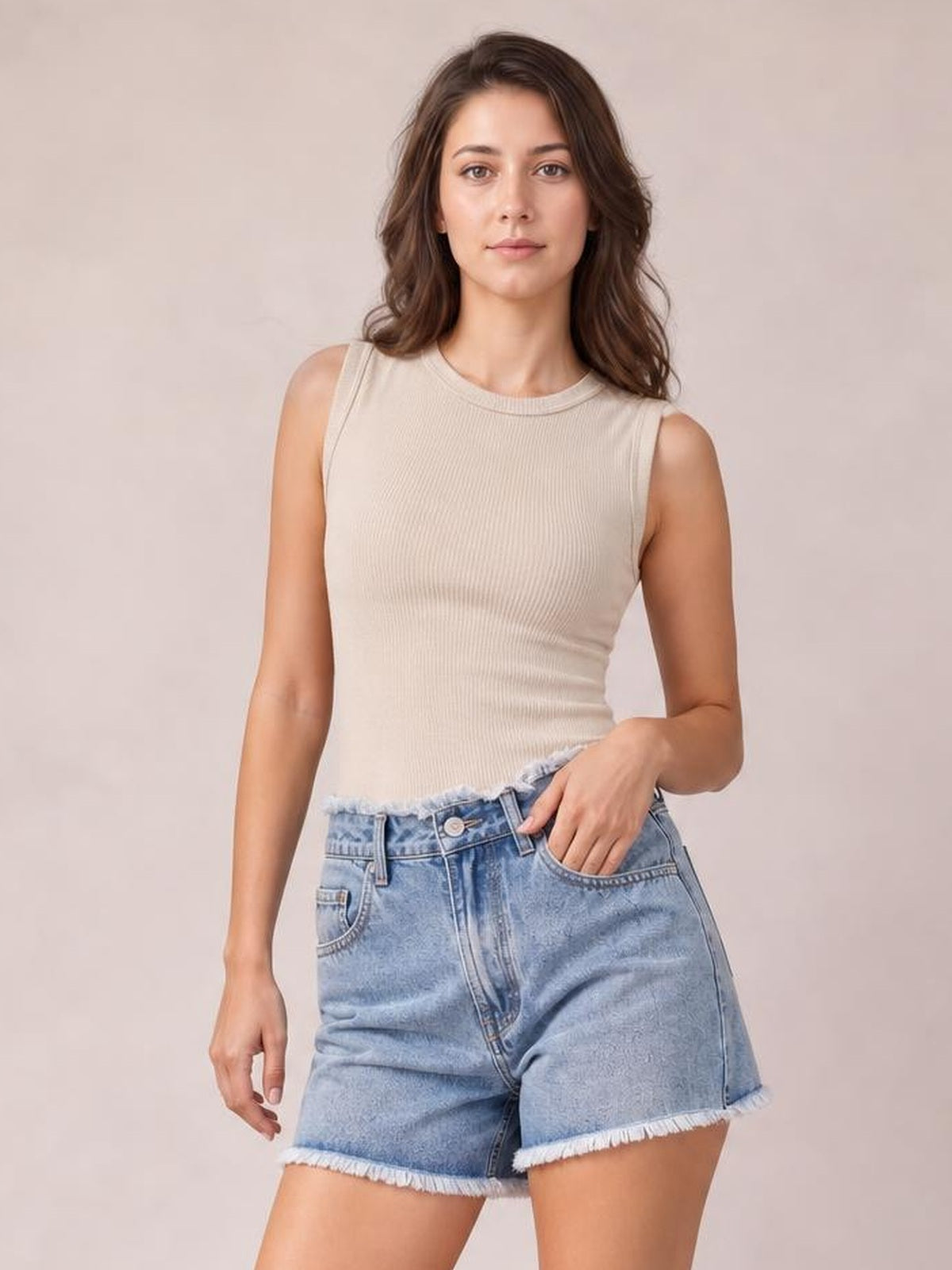 A woman modeling a sleeveless ribbed beige tank top and light-wash denim shorts with frayed hems, standing against a plain beige studio background.
