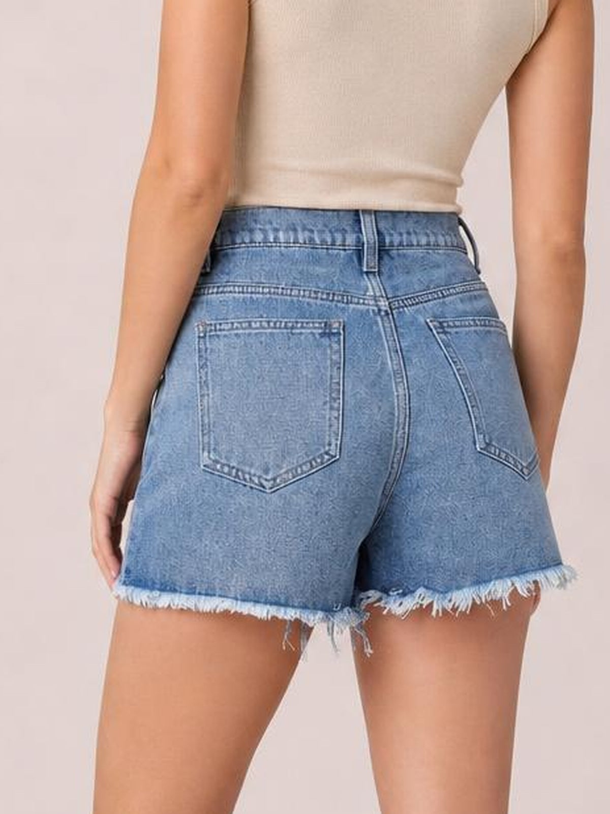 A model poses from behind wearing washed blue denim shorts with a fitted silhouette, belt loops, two back pockets, and frayed raw hems.