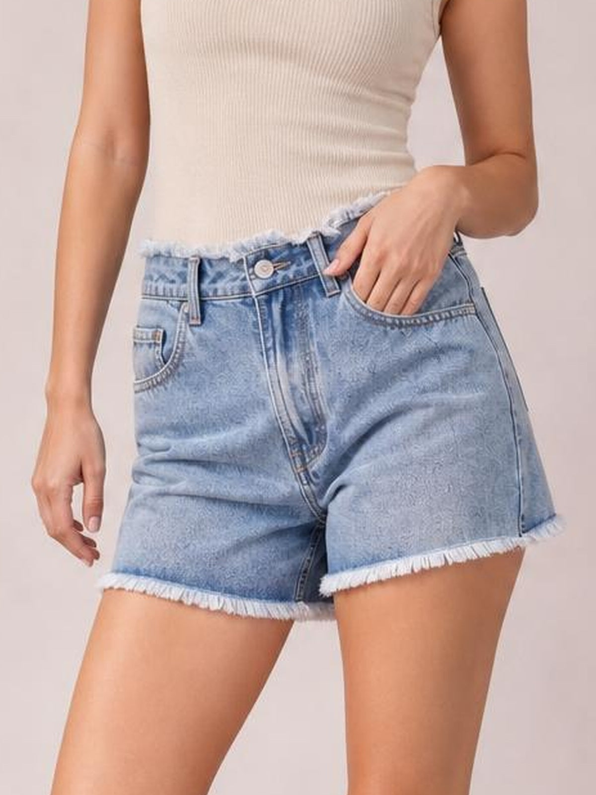 A model wears light blue washed denim shorts with a high-rise fit, five-pocket construction, button closure, belt loops, and frayed raw hems, posing with one hand in a pocket.