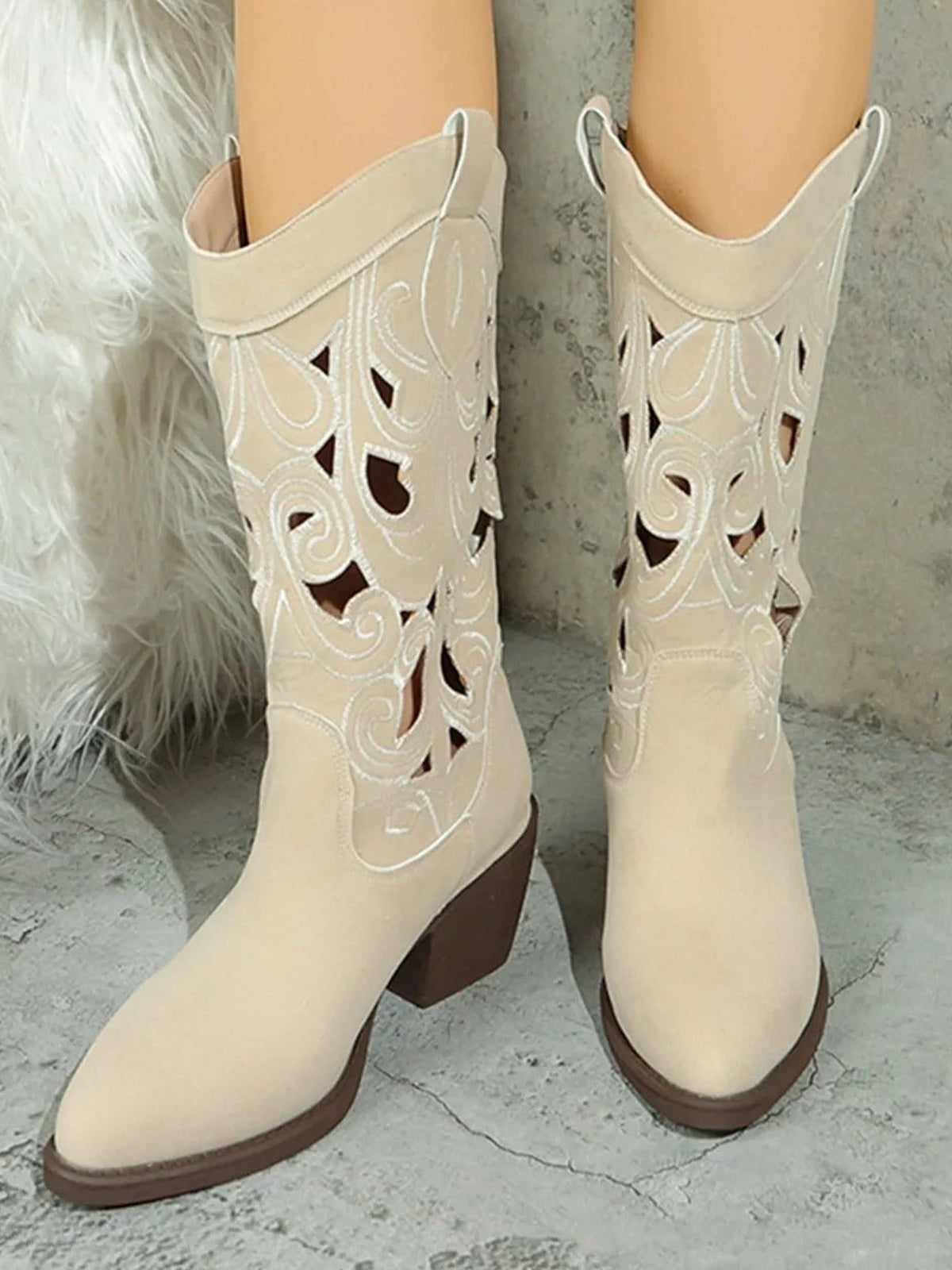 Cream Western boots with pointed toes, mid block heels, and ornate cutout scroll patterns are shown on a model’s feet in a standing pose.