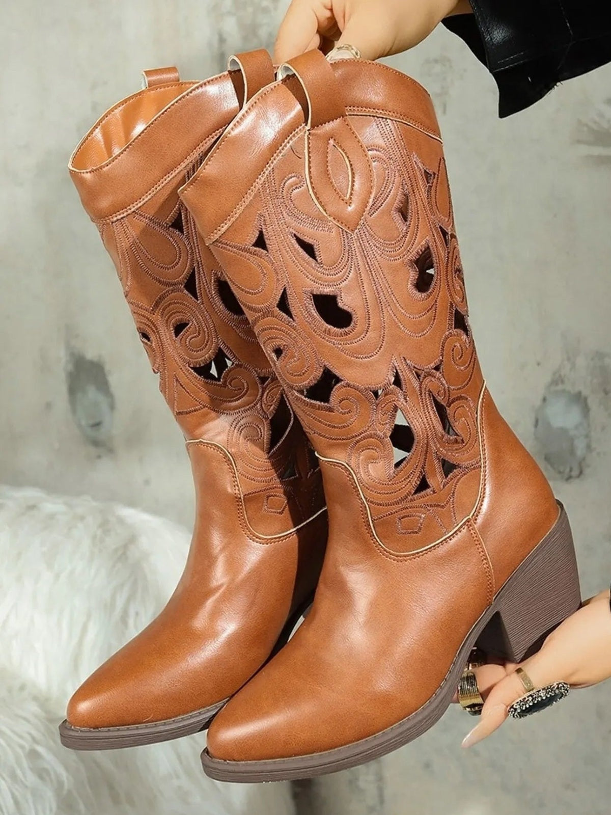 A pair of tan Western boots features pointed toes, mid-calf shafts with ornate cutout patterns, pull tabs, and sturdy mid-height block heels.
