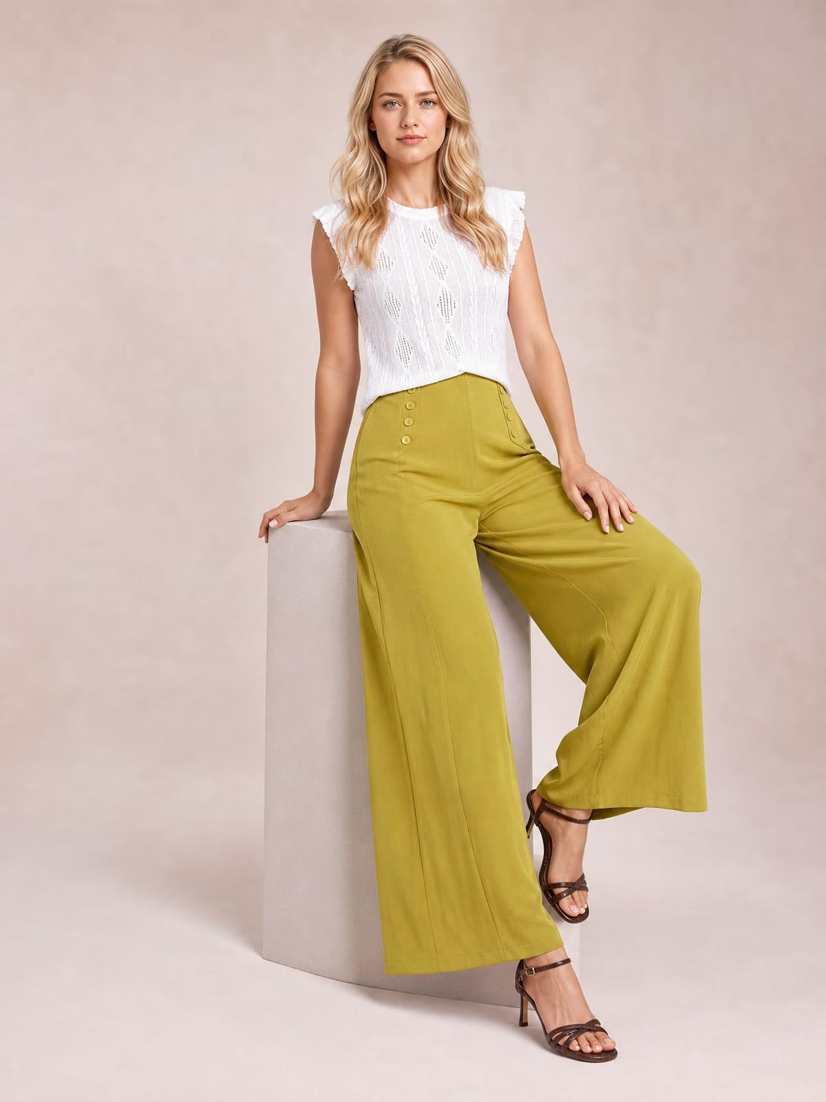 Model seated on a cube wears mustard-green wide-leg pants with decorative buttons, a high waist, front seams, and a flowing full-length silhouette.