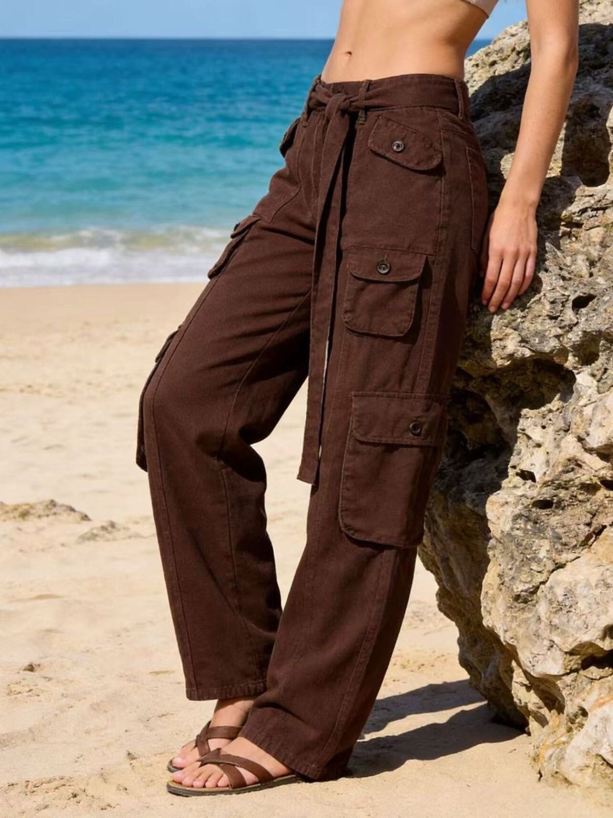 Model leaning against a rock wears brown wide-leg cargo jeans with multiple flap pockets, a tied waist belt, and a relaxed full-length silhouette.