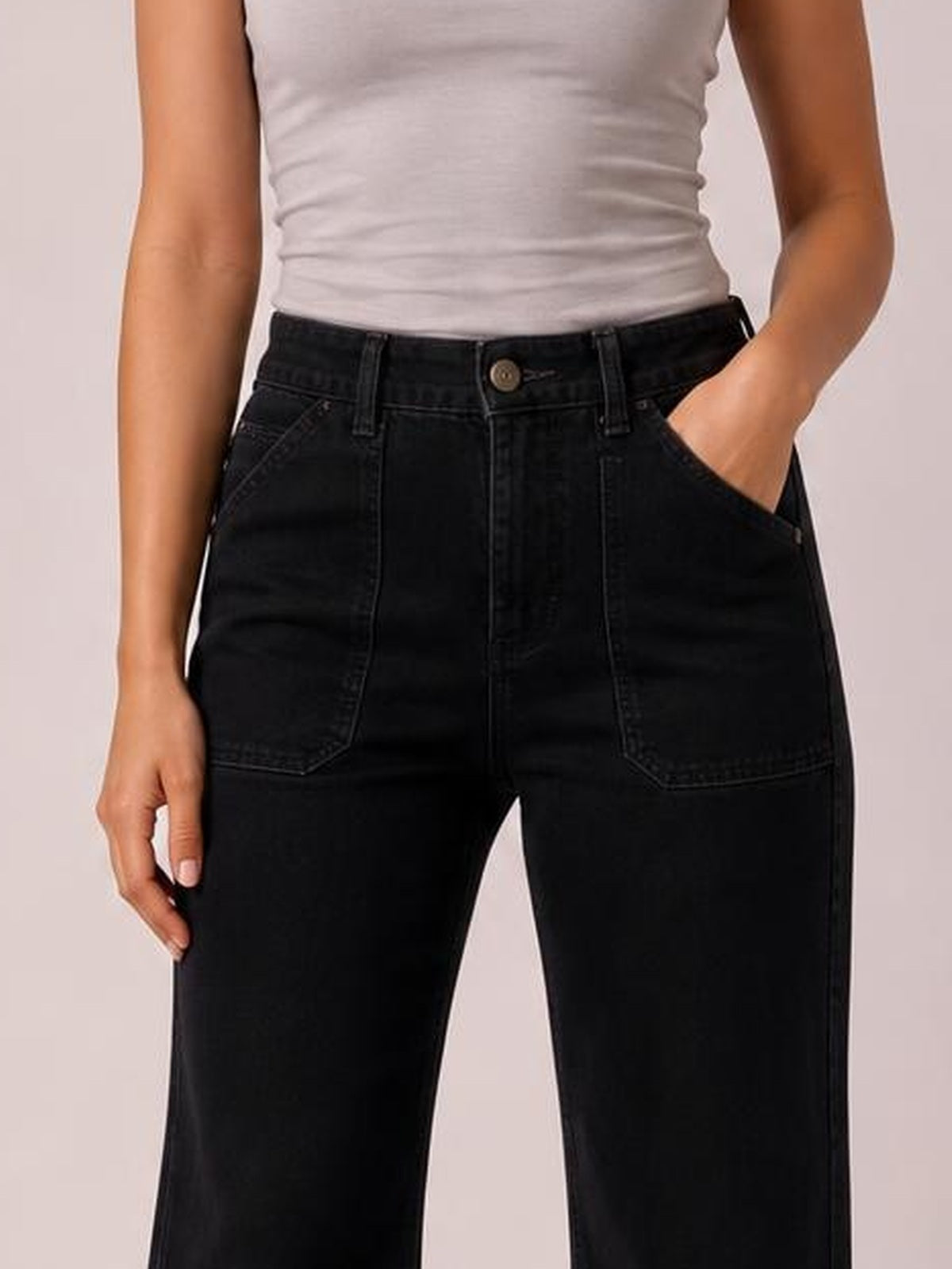 A model wears black wide-leg jeans with contrast stitching, a structured waistband, belt loops, button closure, and large front pockets, posing with one hand tucked inside.