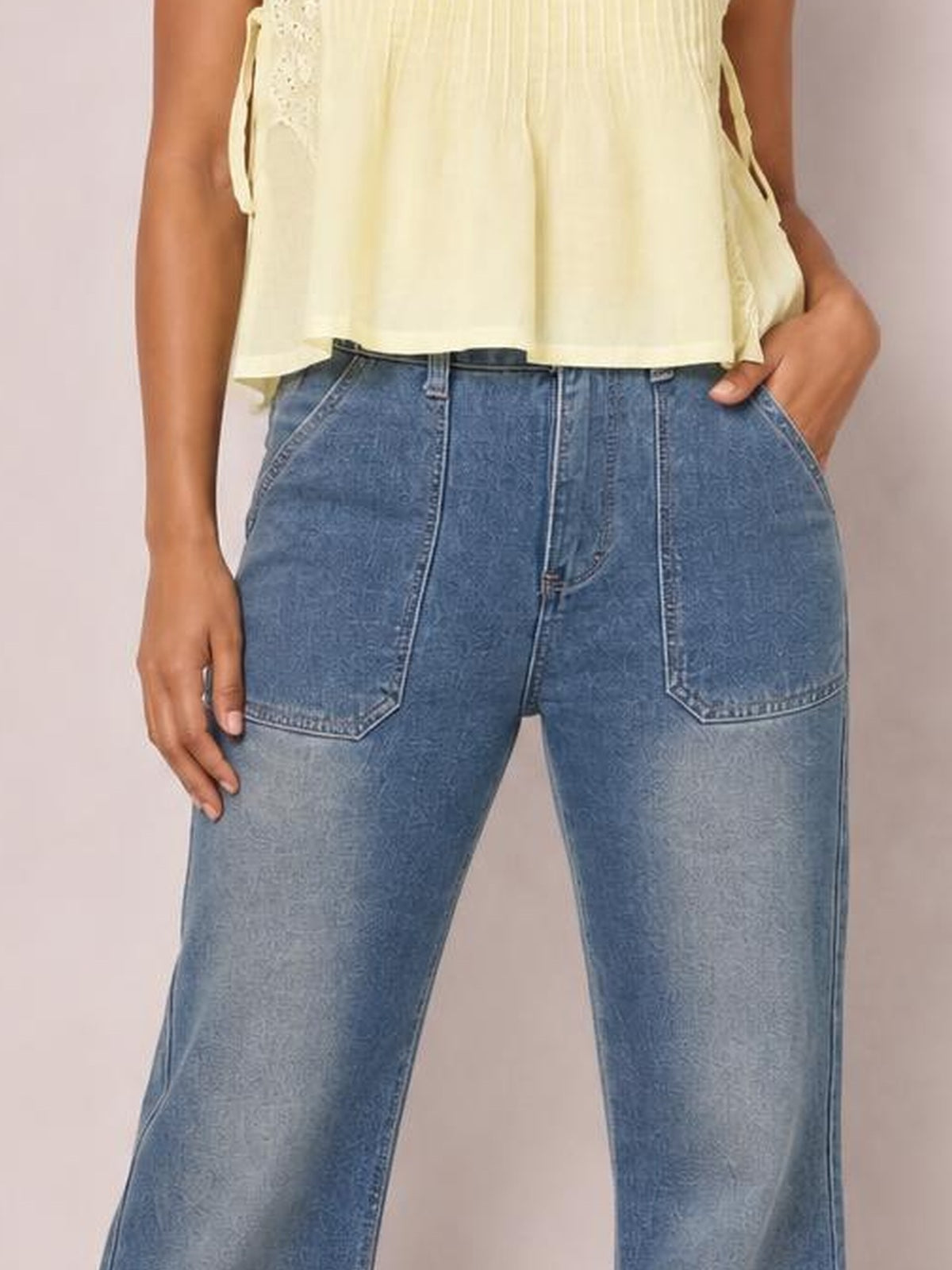 Model wearing blue wide-leg jeans with contrast stitching, a button closure, belt loops, and large front patch pockets, posing with one hand in a pocket.
