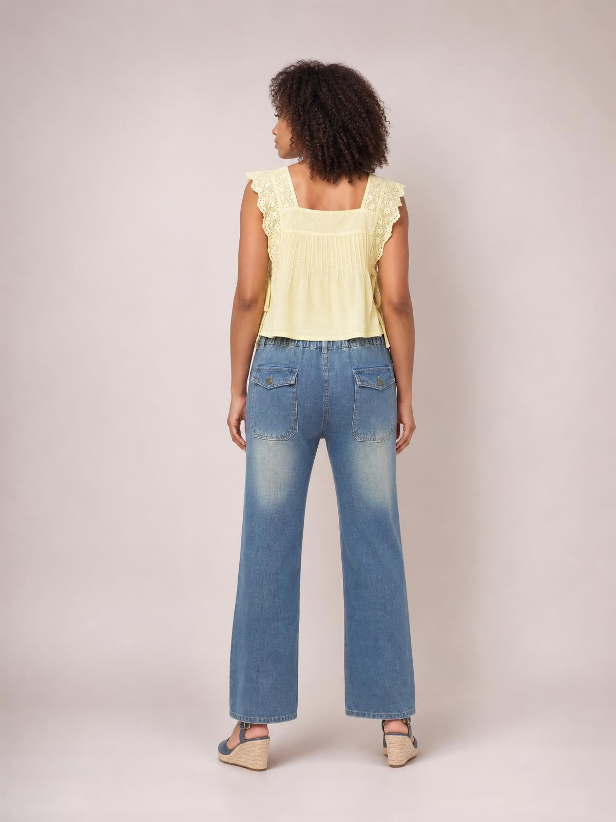 A model stands facing away in light blue wide leg jeans with contrast stitching, belt loops, a buttoned waistband, and two large back patch pockets.