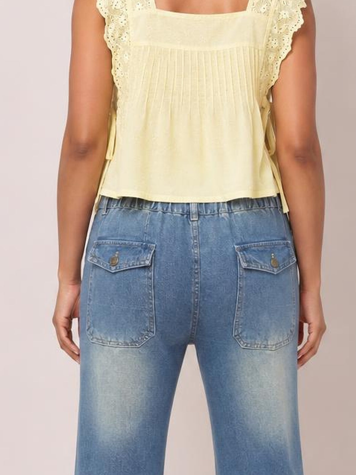 A model stands facing away in light-wash blue wide-leg jeans with visible contrast stitching, a relaxed silhouette, belt loops, and two large back patch pockets.