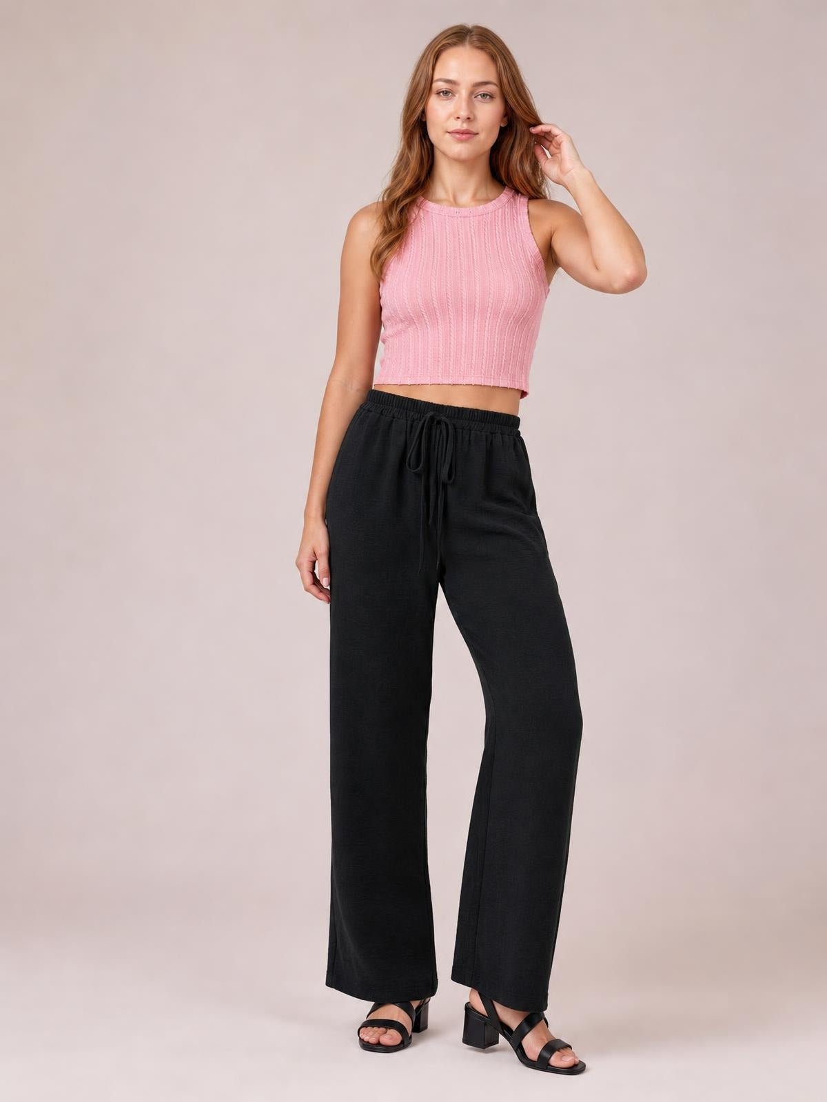 Model poses in black wide leg drawstring pants with an elasticized waist and flowing, relaxed silhouette, styled with a pink sleeveless cropped top.