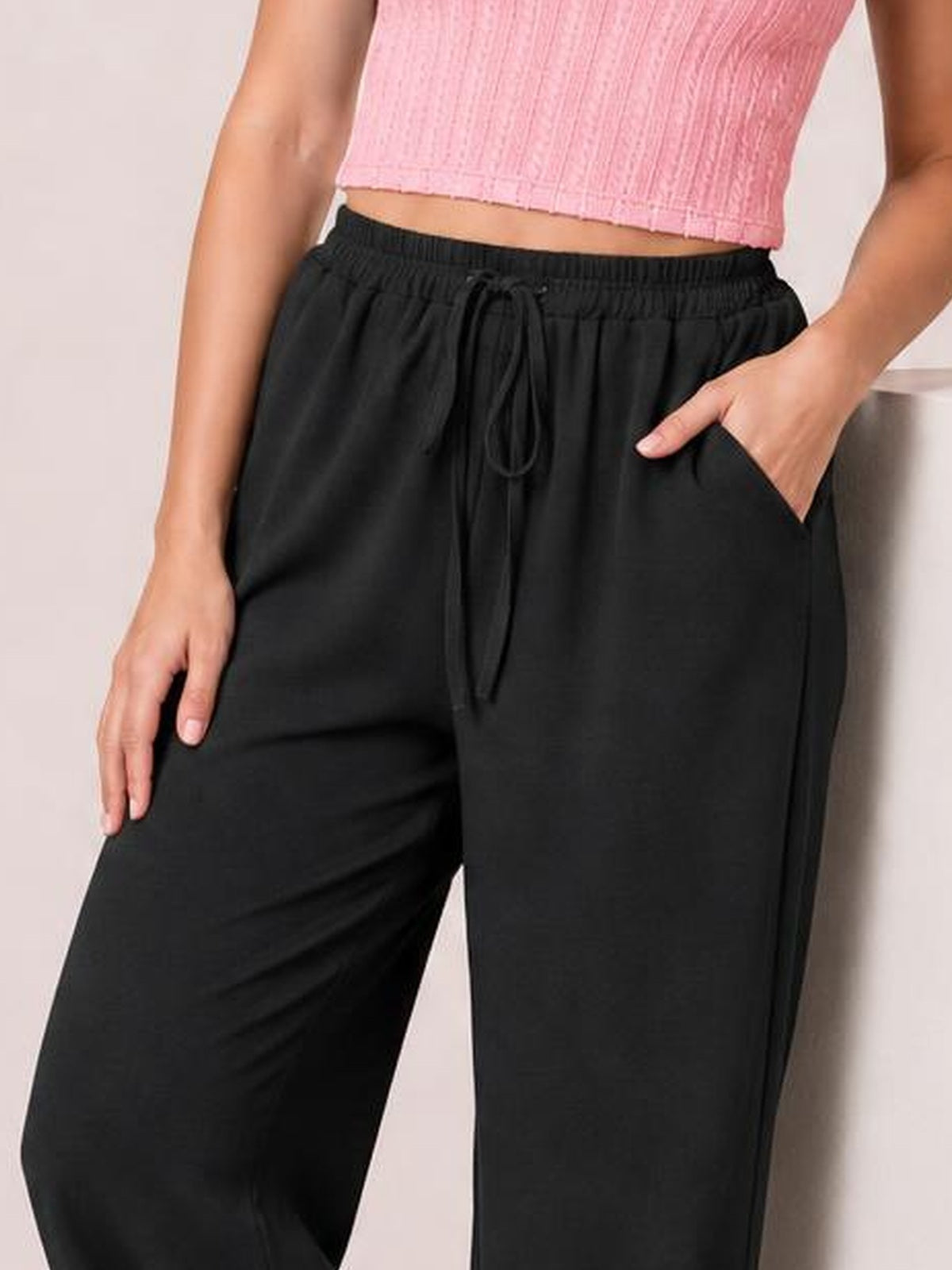 Model wears black wide leg drawstring pants with an elastic waistband, front tie, and side pockets, posing with one hand tucked into a pocket.