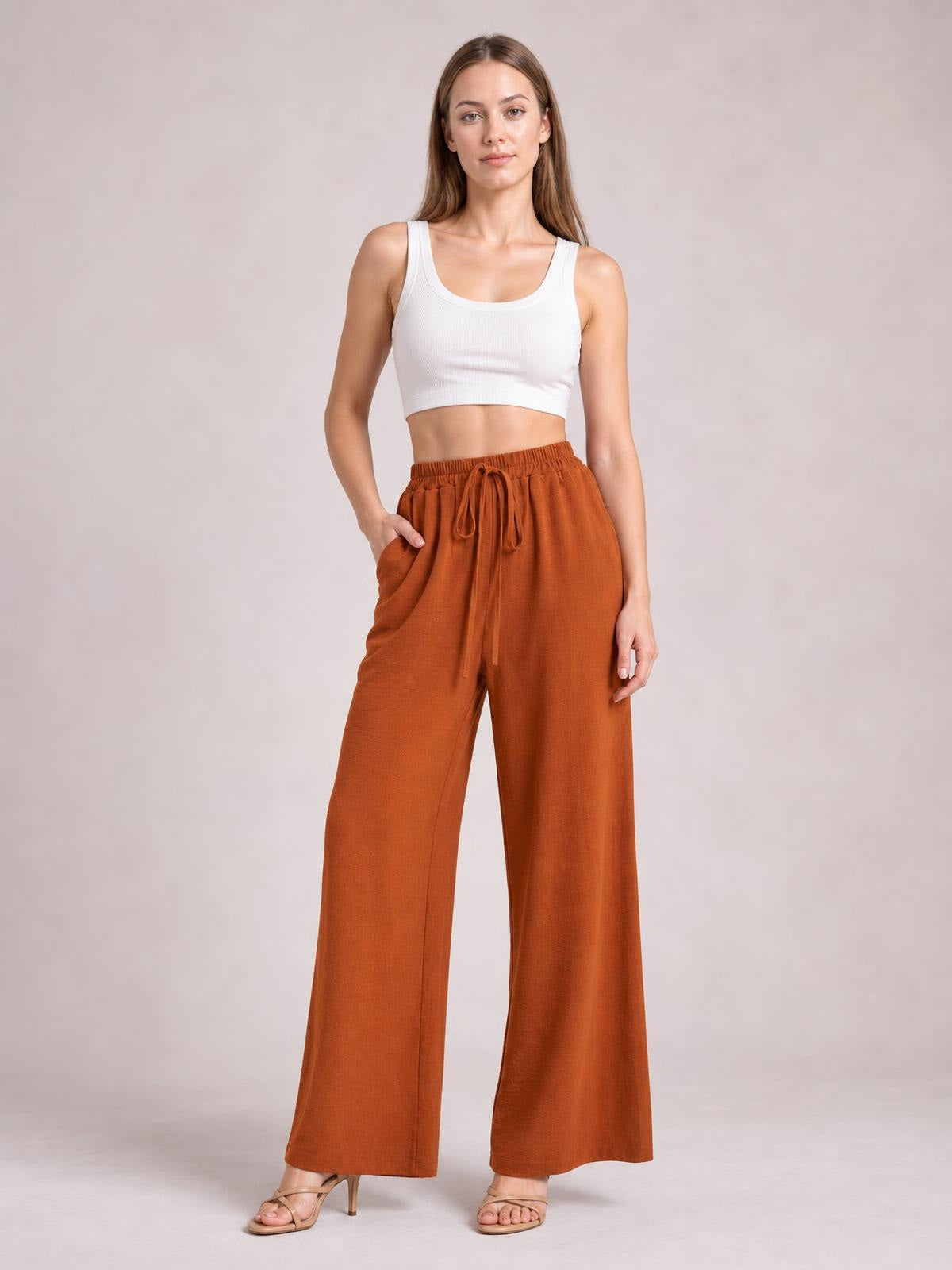 A model poses in rust-colored wide leg drawstring pants with an elastic waist, front tie, side pockets, and a relaxed flowy silhouette.