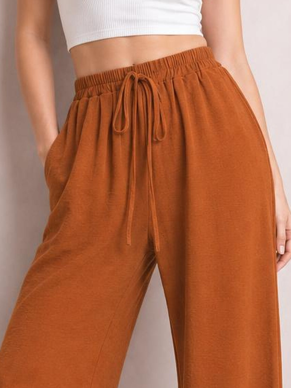 Model shown from the waist down wears rust-orange wide-leg drawstring pants with an elastic waist, front tie, side pockets, and a flowy silhouette.
