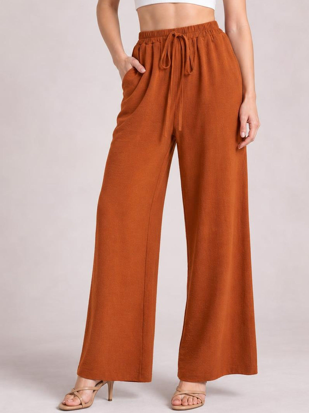 Model wearing rust-orange wide leg drawstring pants with an elastic waist, tied drawstring, flowing silhouette, and hands resting in the side pockets.
