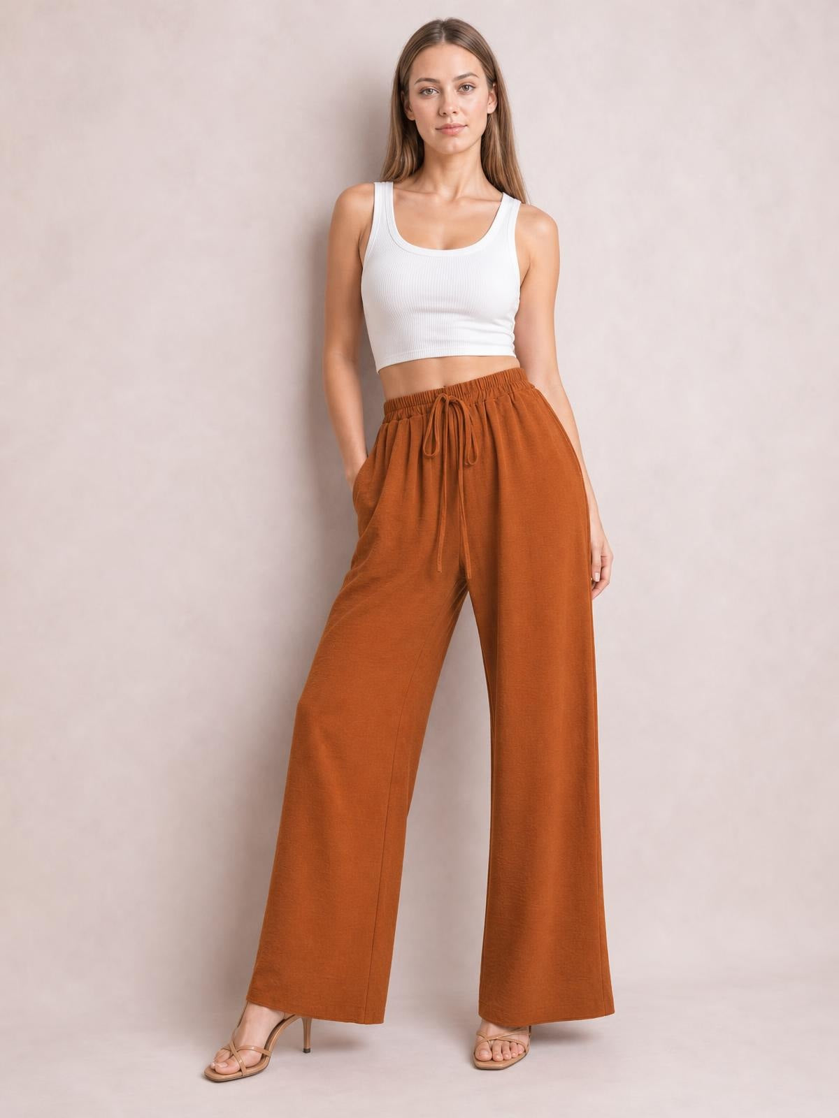 Model poses in rust-brown wide-leg drawstring pants with an elasticized waist, front tie, and relaxed flowy silhouette.