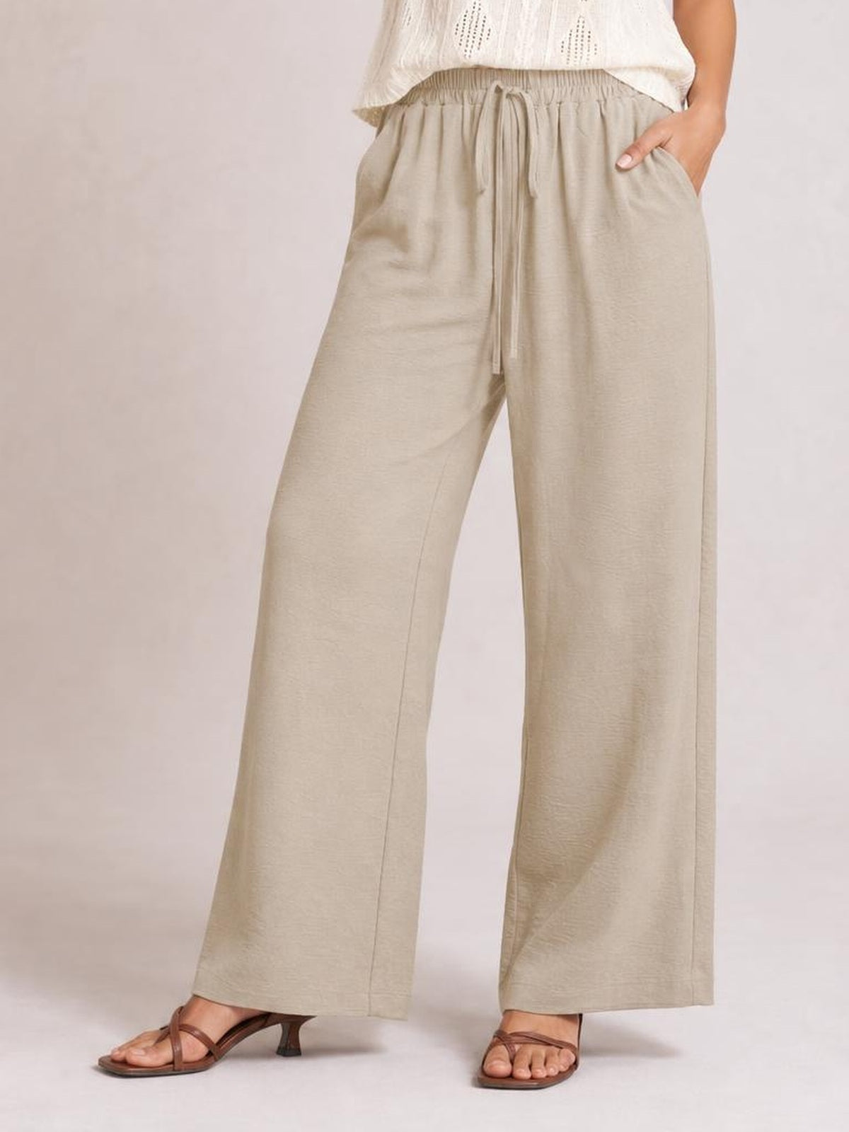 Model wearing beige wide leg drawstring pants with a relaxed flowy silhouette, elastic waist, front tie, side pockets, and straight-leg construction.