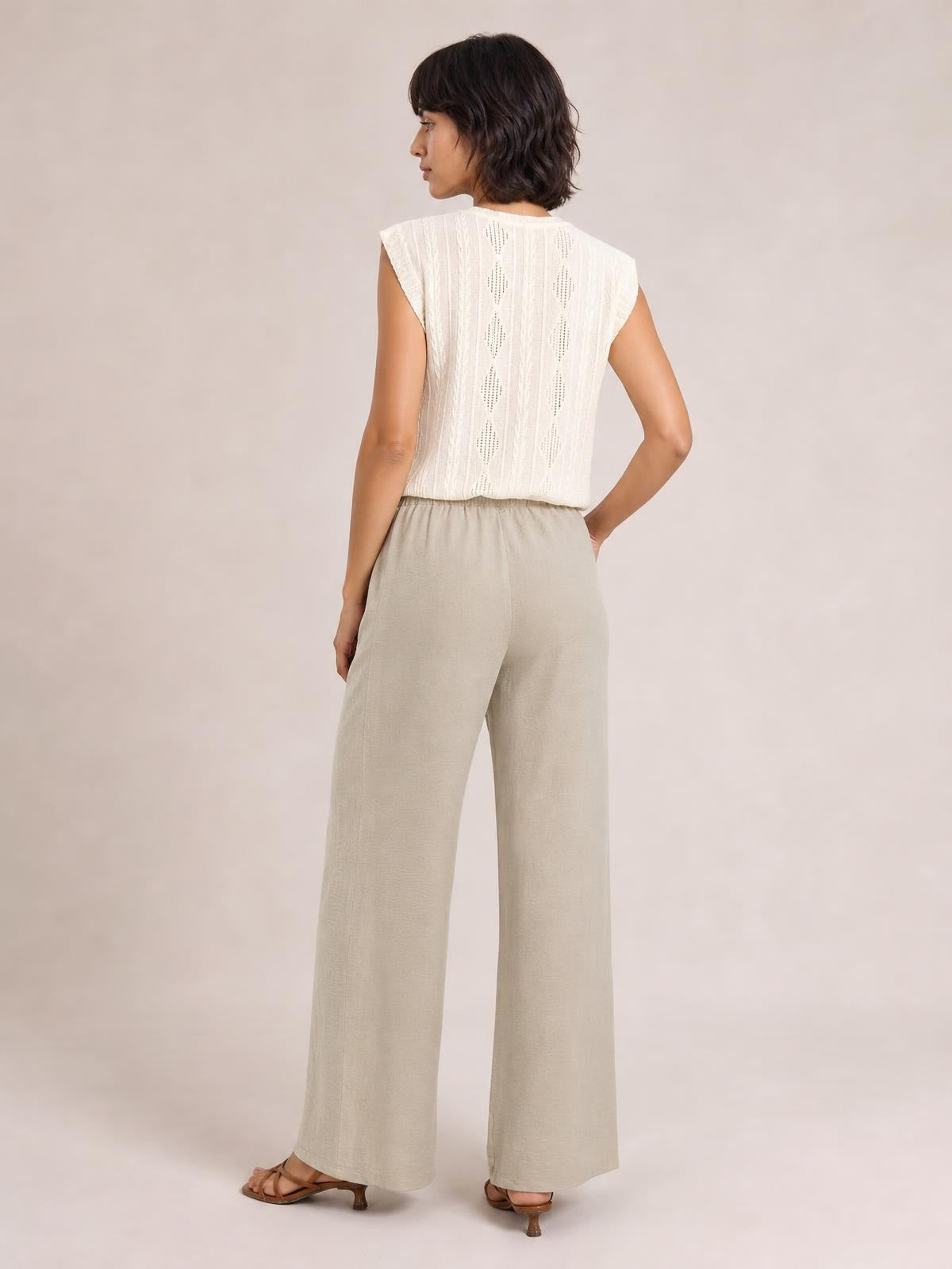 A model poses in profile from the back wearing beige wide-leg drawstring pants with a relaxed, flowing silhouette, elasticized waist, and full-length hems.