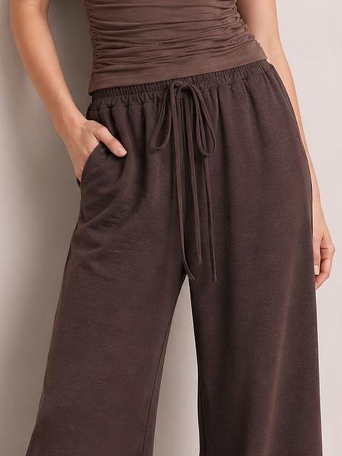 Model wearing dark brown wide leg drawstring pants with an elasticized waistband, long front drawstrings, side pockets, and a relaxed flowy silhouette.