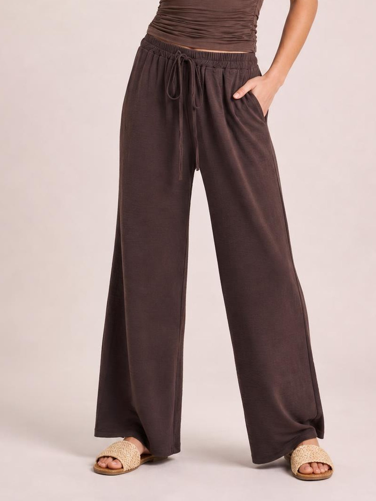 Model wears dark brown wide-leg drawstring pants with an elastic waistband, long flowy legs, and one hand tucked into a side pocket.