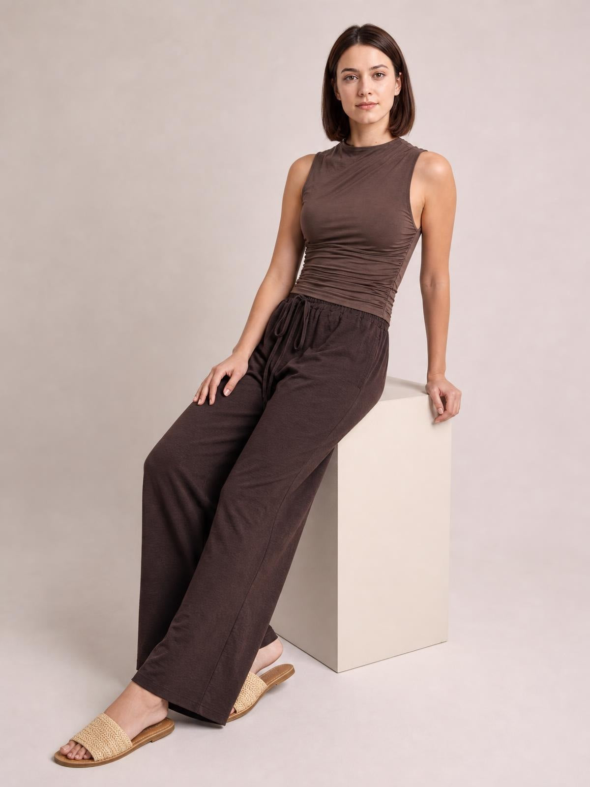 A model leans on a white block wearing dark brown wide-leg drawstring pants with a relaxed, flowy silhouette and full-length hems.