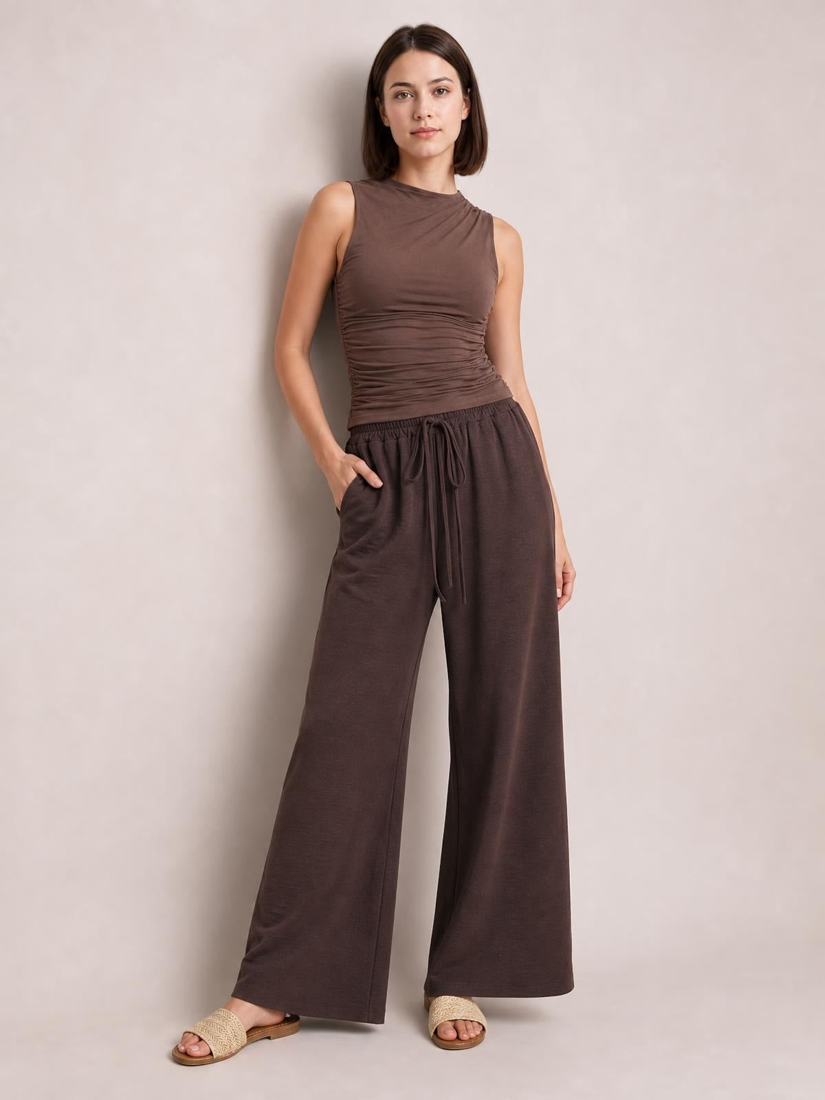 A model poses in dark brown wide leg drawstring pants with a relaxed full-length silhouette, elastic waist, front tie, and side pockets.
